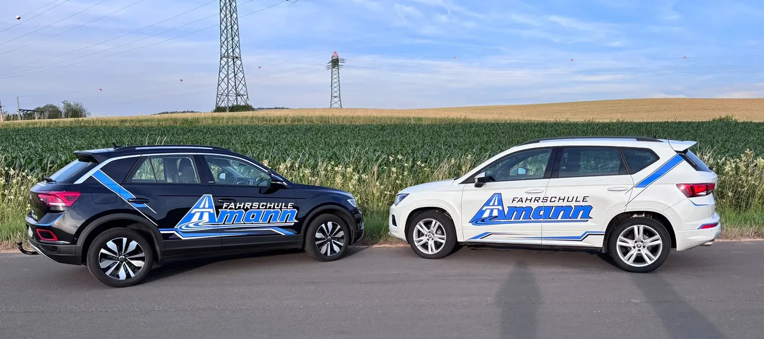 Amann - Fahrschule – Das Bild zeigt zwei Fahrzeuge mit der Aufschrift „Fahrschule Amann“. Der Wagen links ist ein schwarzer Volkswagen T-Roc, der SUV rechts ein weißer SEAT Ateca. Beide sind wahrscheinlich mit Doppelsteuerungssystemen ausgestattet, wie sie für die Fahrausbildung üblich sind.